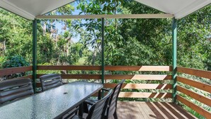 Une terrasse extérieure couverte, dotée d’une table et de chaises en verre, entourée de rampes en bois et d’une végétation luxuriante.