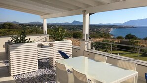 Une terrasse avec une table et des chaises blanches, un tapis à motifs et une vue sur un paysage côtier avec des montagnes.