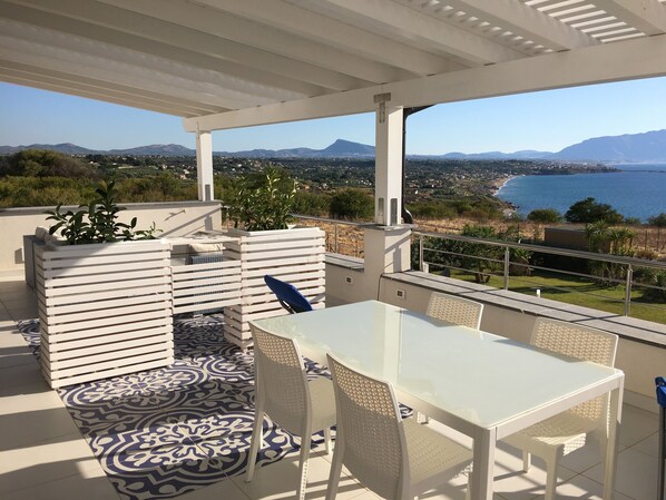 Eine Terrasse mit einem weißen Tisch und Stühlen, einem gemusterten Teppich und Blick auf eine Küstenlandschaft mit Bergen.