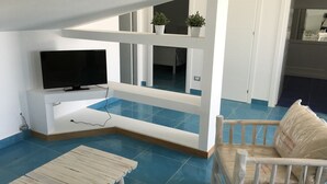 Un salon avec un téléviseur posé sur un meuble blanc, un banc en bois, une chaise blanche et un sol carrelé de carreaux bleus.