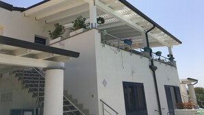 Un bâtiment à la façade blanche, doté d’un balcon couvert orné de plantes en pot et d’un escalier menant à un étage supérieur.