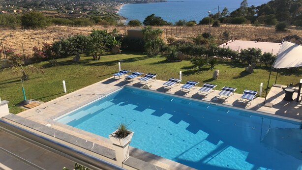 Une piscine entourée de chaises longues, bordée d’une pelouse et offrant une vue sur la mer et les montagnes.