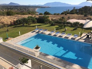 Ein Pool mit Liegestühlen, umgeben von einer Rasenfläche und Blick auf das Meer und die Berge.