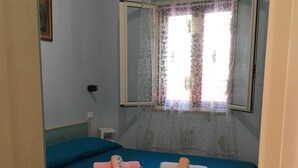 A room with a bed covered in a teal bedspread, two rolled towels on the bed, a window with a floral curtain, and a mirror above the bed. - MIMOSA A Three-room detached house in the Villa Carla Residence for realiz (Vieste)