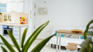 Ein modernes, minimalistisches Zimmer mit Küchenzeile, weißen Schränken und einer Arbeitsplatte mit Obst. Ein Bett mit Holz-Kopfteil und ein kleiner Tisch mit einer Schüssel darauf.