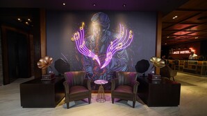 A lobby with a neon mural, leather chairs, a small round table, and a reception desk. - Jazz Hotel Penang (George Town)