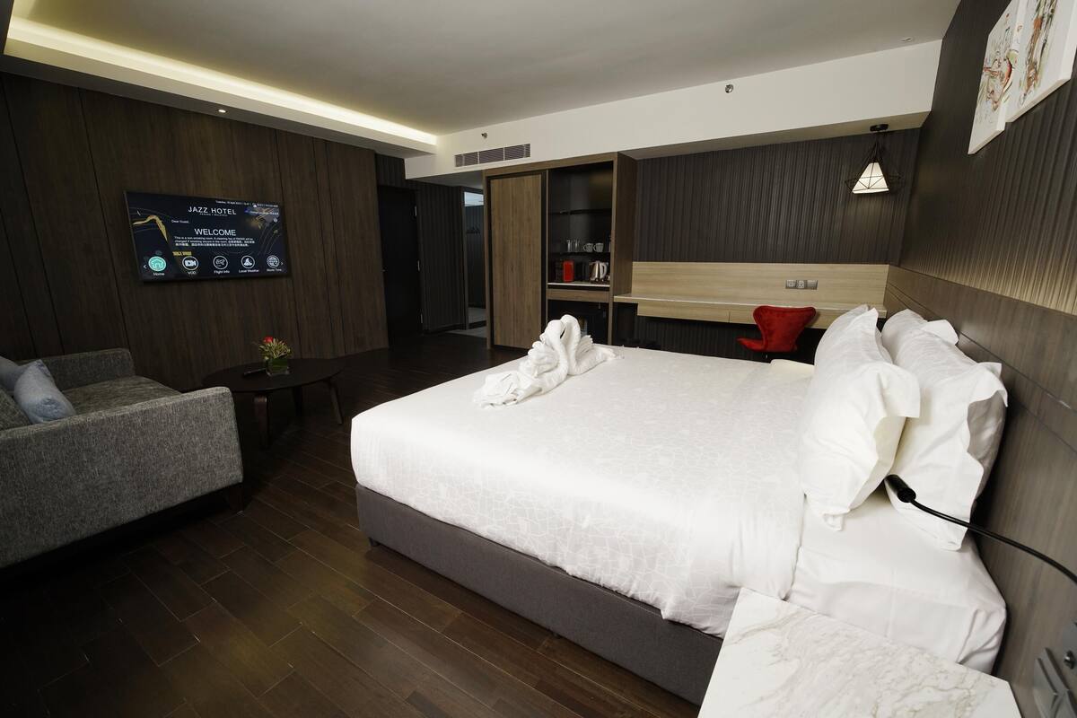 Ett modernt hotellrum med en stor säng, en sittgrupp, en platt-TV som visar hotellets välkomstmeddelande och en garderob i trä.