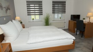 A neatly made bed in a room with a television, desk, and two windows with blinds. - Hotel Garni - Dahlbrucher Hof (Hilchenbach)