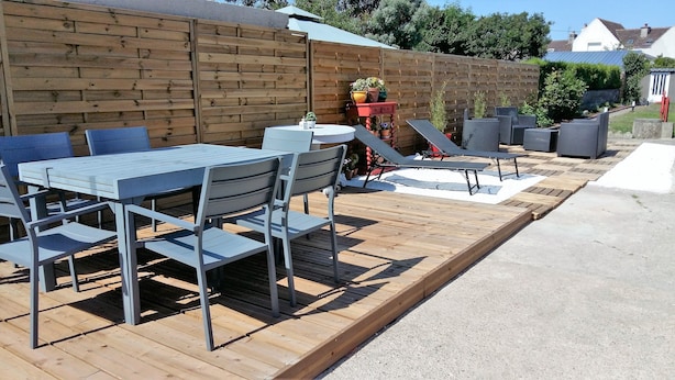 Un patio exterior con terraza de madera, una mesa con sillas y una pequeña mesa redonda con una planta en maceta.