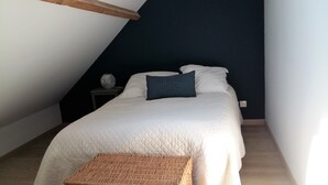Un dormitorio con techo inclinado, una cama con ropa de cama blanca, un almohadón azul oscuro y una pequeña mesa redonda con un jarrón.