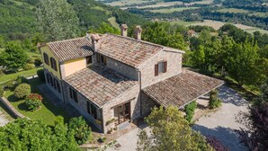 Une maison en pierre avec un toit de tuiles, entourée d’une végétation luxuriante et de collines.