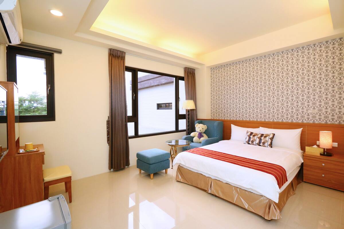 Une chambre d’hôtel avec un grand lit, un bureau avec une chaise, une petite table et un ours en peluche.