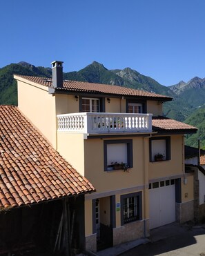 Un edificio de dos pisos con balcón, tejado de tejas y un paisaje montañoso al fondo.