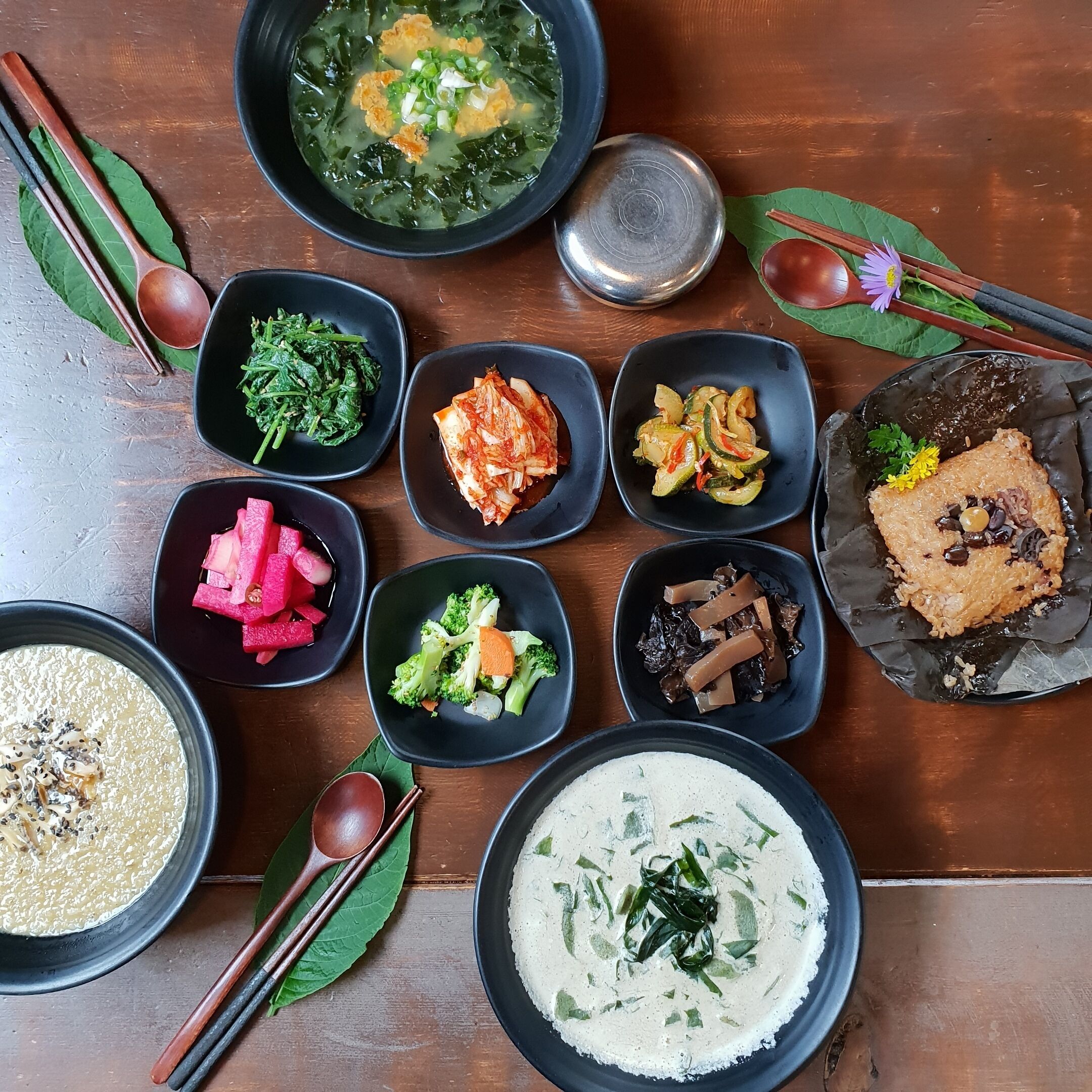 A variety of dishes arranged on a wooden table, including a bowl of soup, several side dishes, and a plate of rice.