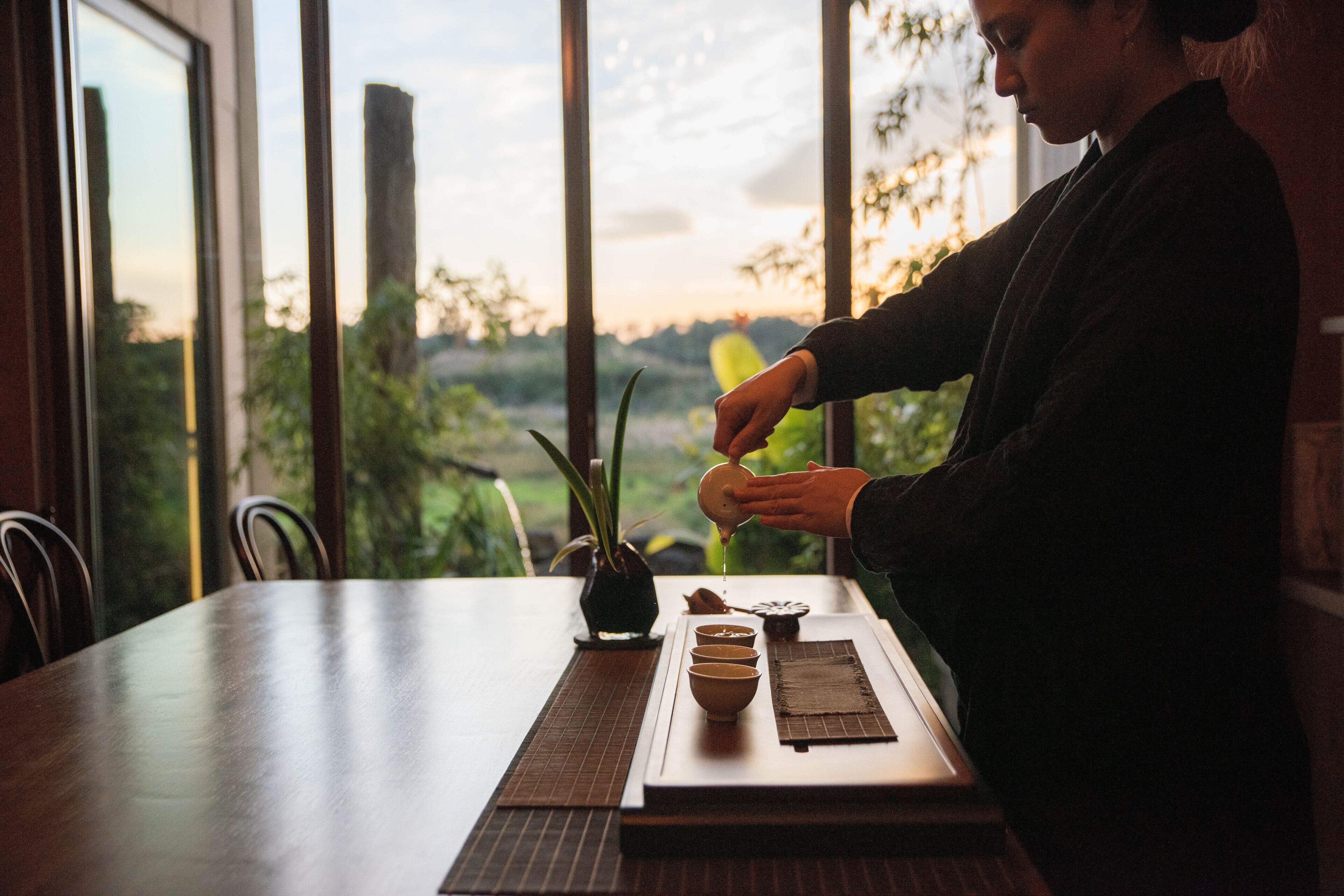 A person pouring tea into a cup at a table with a view of greenery and buildings.