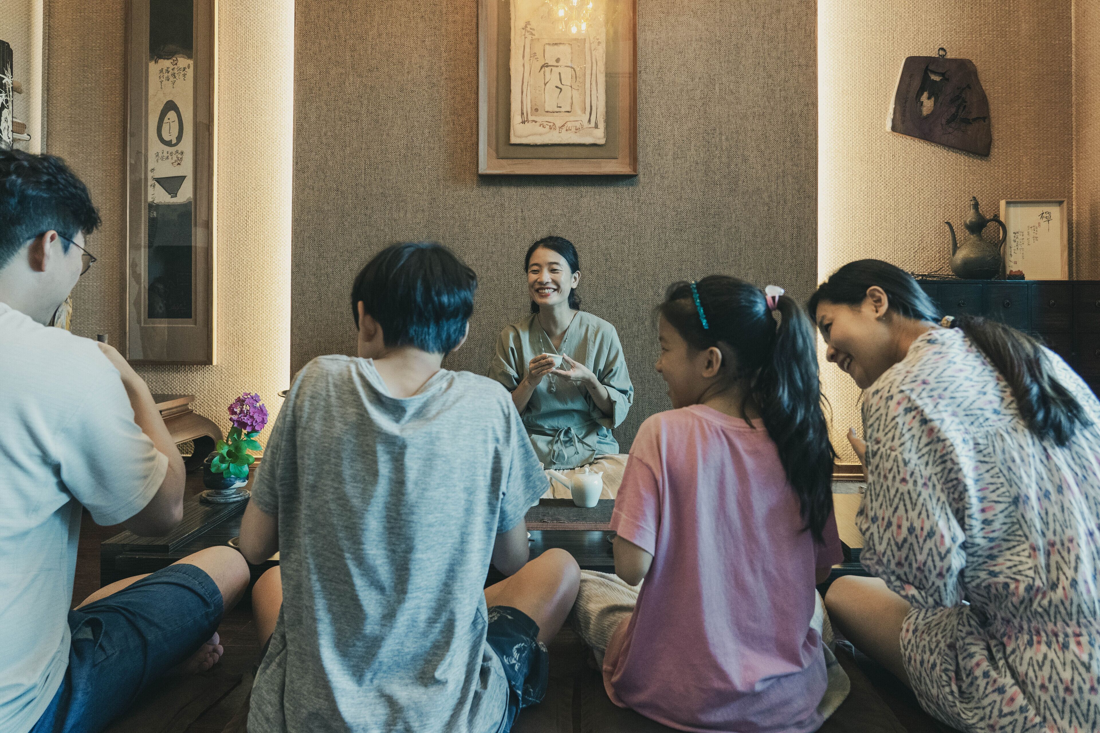 A group of people sitting on the floor in a room with a wooden floor, a framed picture on the wall, and a small table with a plant.
