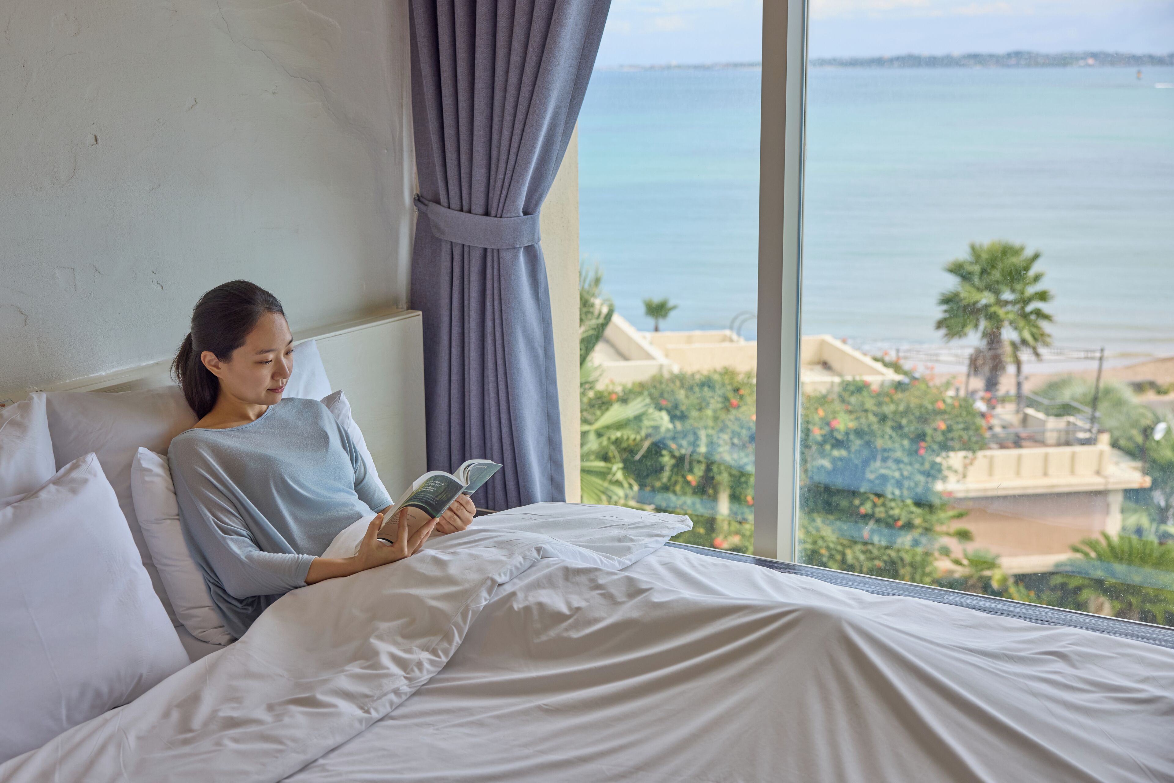 Una persona leyendo un libro en una habitación con un amplio ventanal que da a una playa y palmeras.