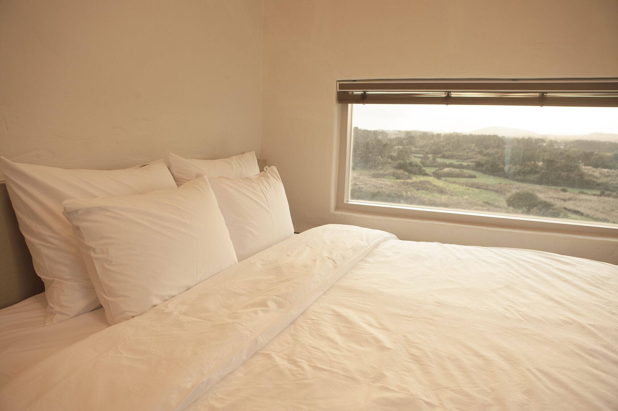 Un letto con biancheria e cuscini bianchi, una finestra vista su un paesaggio e una parete beige.