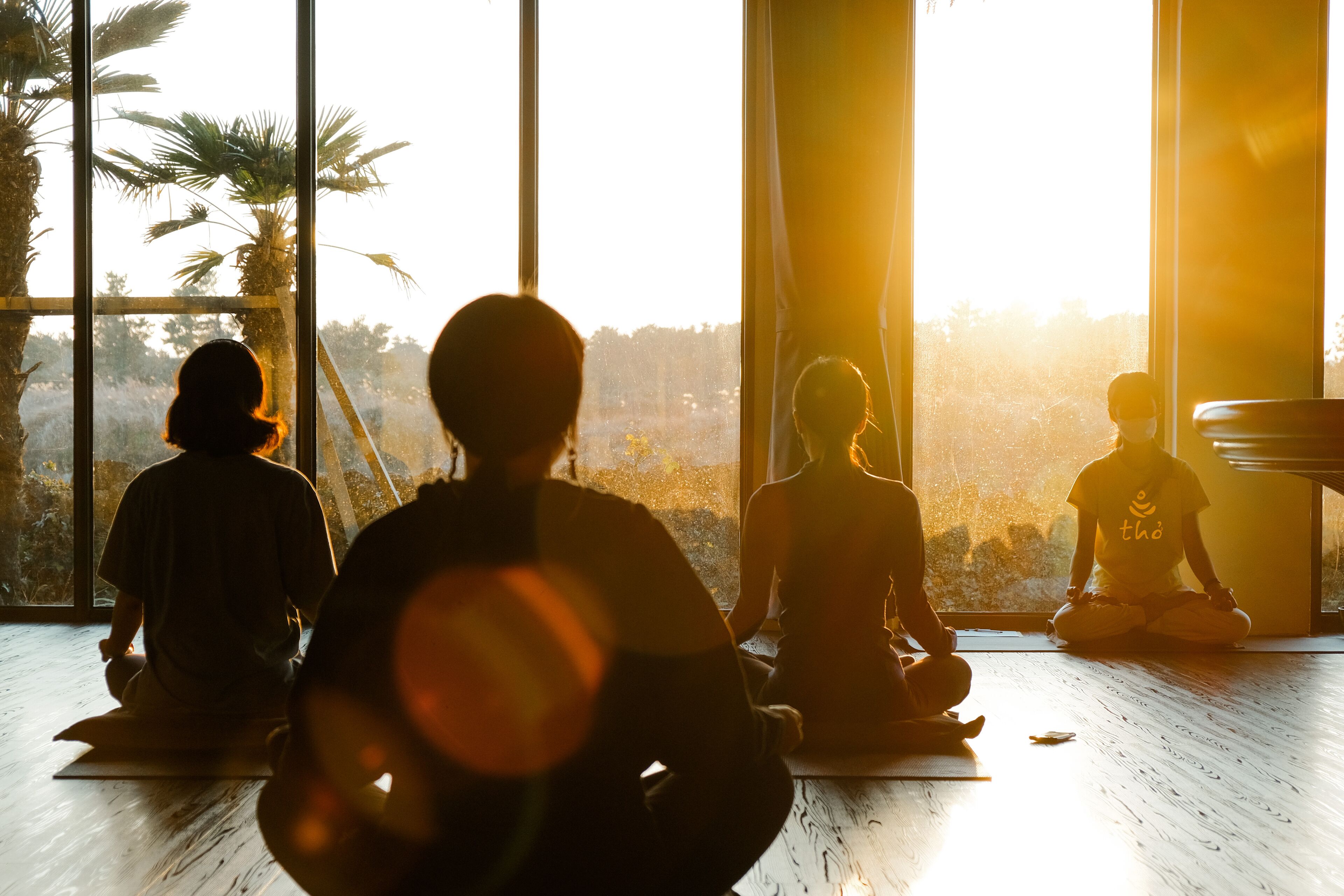 Cuatro personas meditando en una habitación con amplios ventanales, piso de madera y vistas a palmeras.