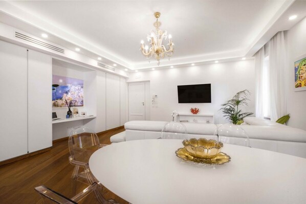Una sala de estar moderna con un sofá blanco, una mesa de comedor redonda con un centro dorado, una araña y un televisor de pantalla plana montado en la pared.