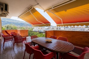 A terrace with a wooden table and red chairs, a brick wall, and a view of buildings and hills.