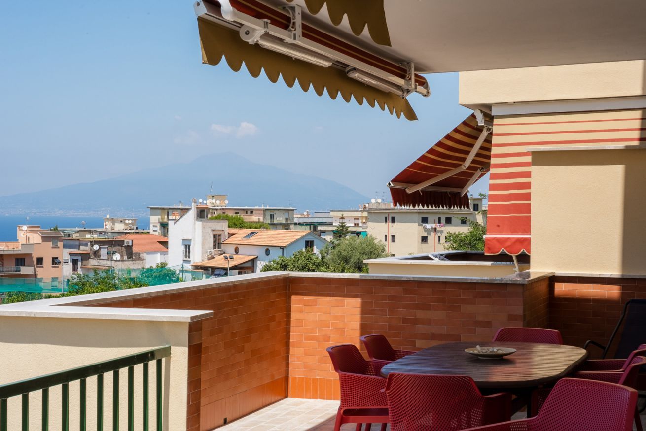A balcony with a table and chairs, a barbecue grill, and a view of a cityscape with buildings and a mountain in the distance.