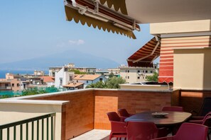 A balcony with a table and chairs, a barbecue grill, and a view of a cityscape with buildings and a mountain in the distance.
