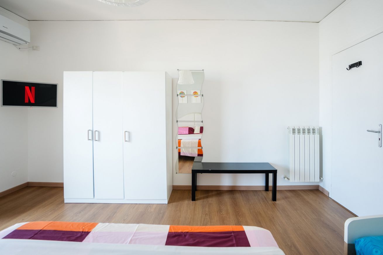 Une chambre avec une armoire, un lit recouvert d’une courtepointe colorée, un banc, un radiateur et un téléviseur affichant le logo de Netflix.