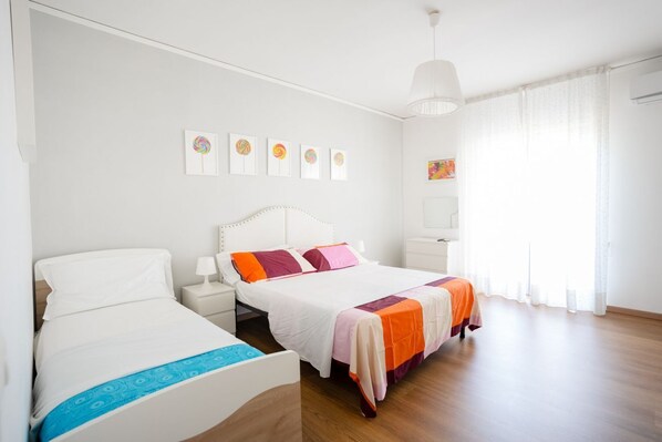 A hotel room with two beds, a white headboard, a bedside table with a lamp, a large window with sheer curtains, and wall art.