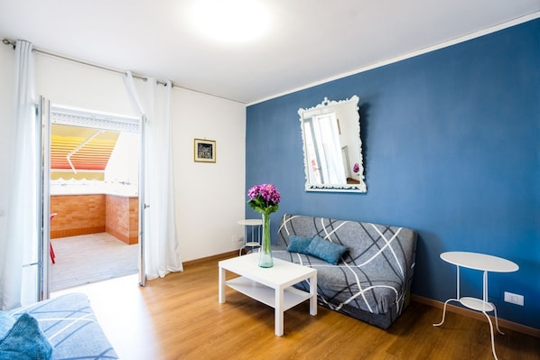 Una habitación con una pared azul, un sofá, una mesita con un jarrón, un espejo y una puerta que da a un balcón.