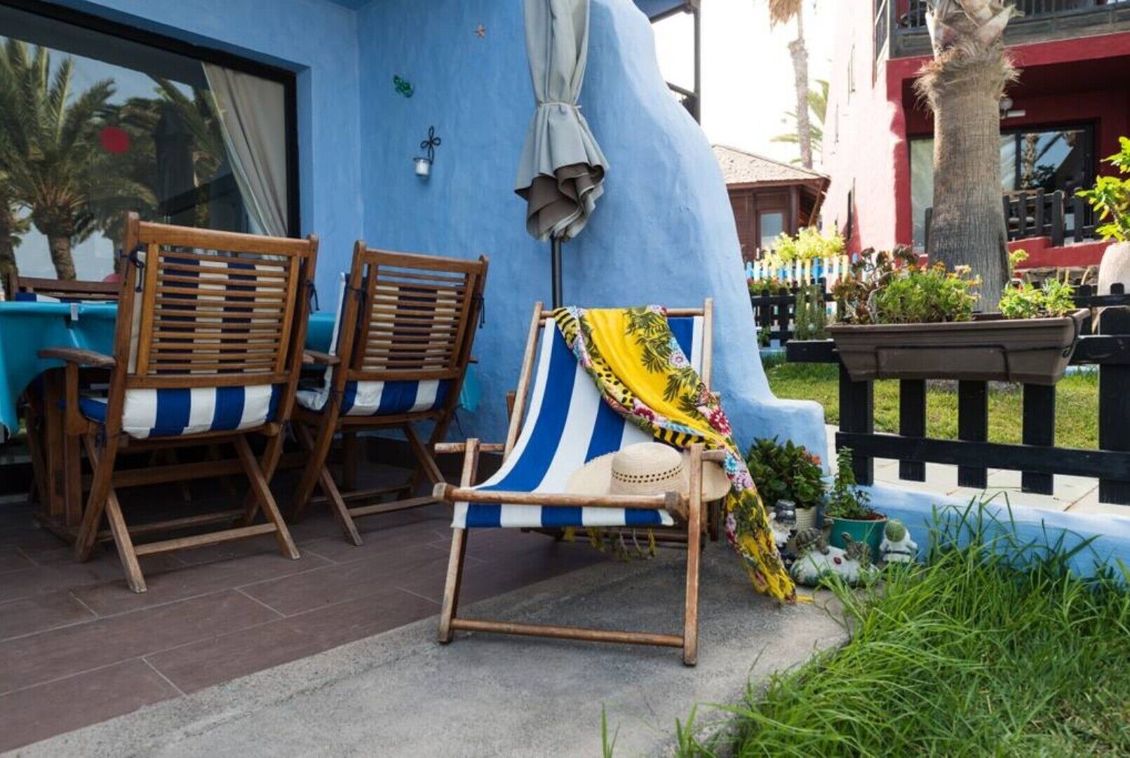 A patio area with two wooden chairs and a striped beach chair, a small table, and a blue wall with a window.