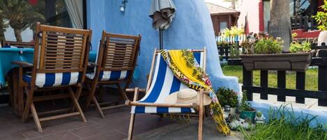A patio area with two wooden chairs and a striped beach chair, a small table, and a blue wall with a window.