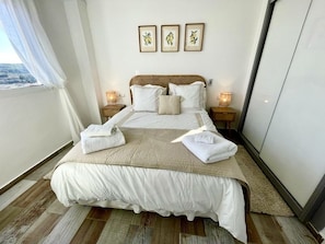 A neatly made bed with white linens and a beige throw blanket, two bedside lamps, and three framed botanical prints on the wall.