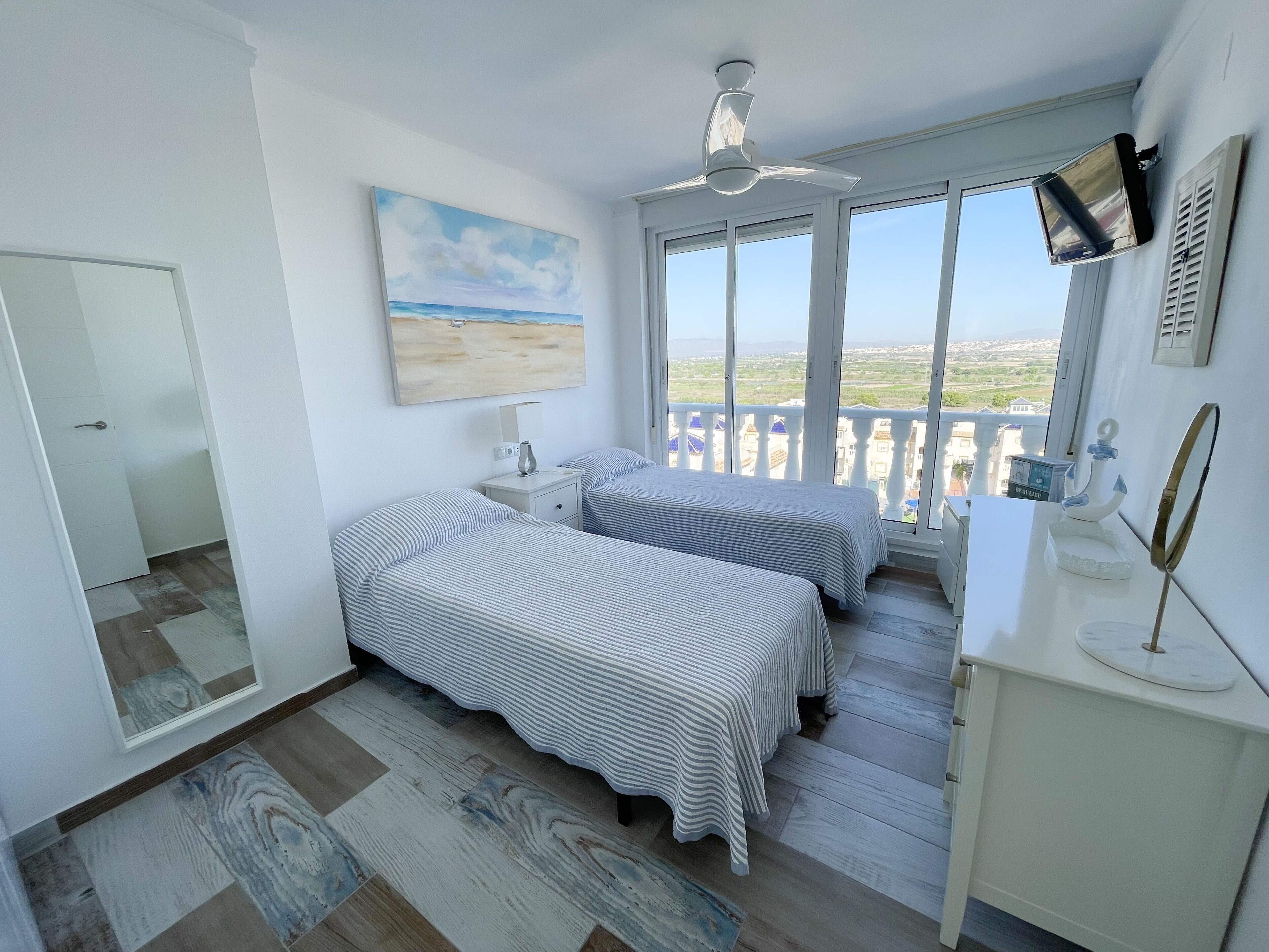 Une chambre avec deux lits, un ventilateur de plafond, un tableau représentant une plage et un balcon offrant une vue sur le paysage.