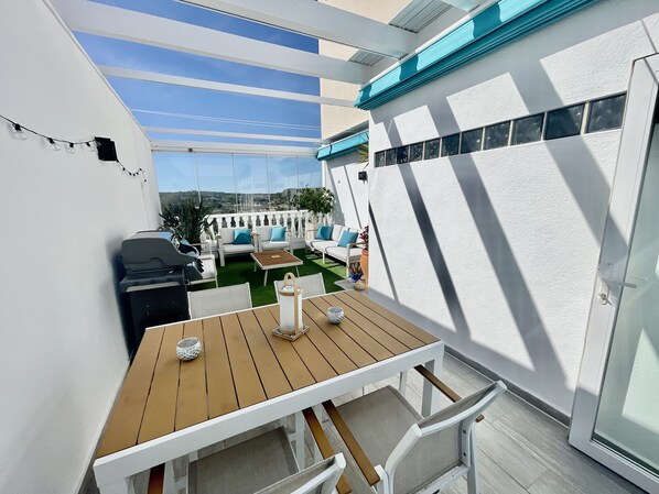 A patio with a wooden table and white chairs, a grill, and a view of the landscape.