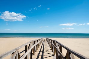 A wooden pier leading to a sandy beach with clear blue water and a few clouds in the sky.
