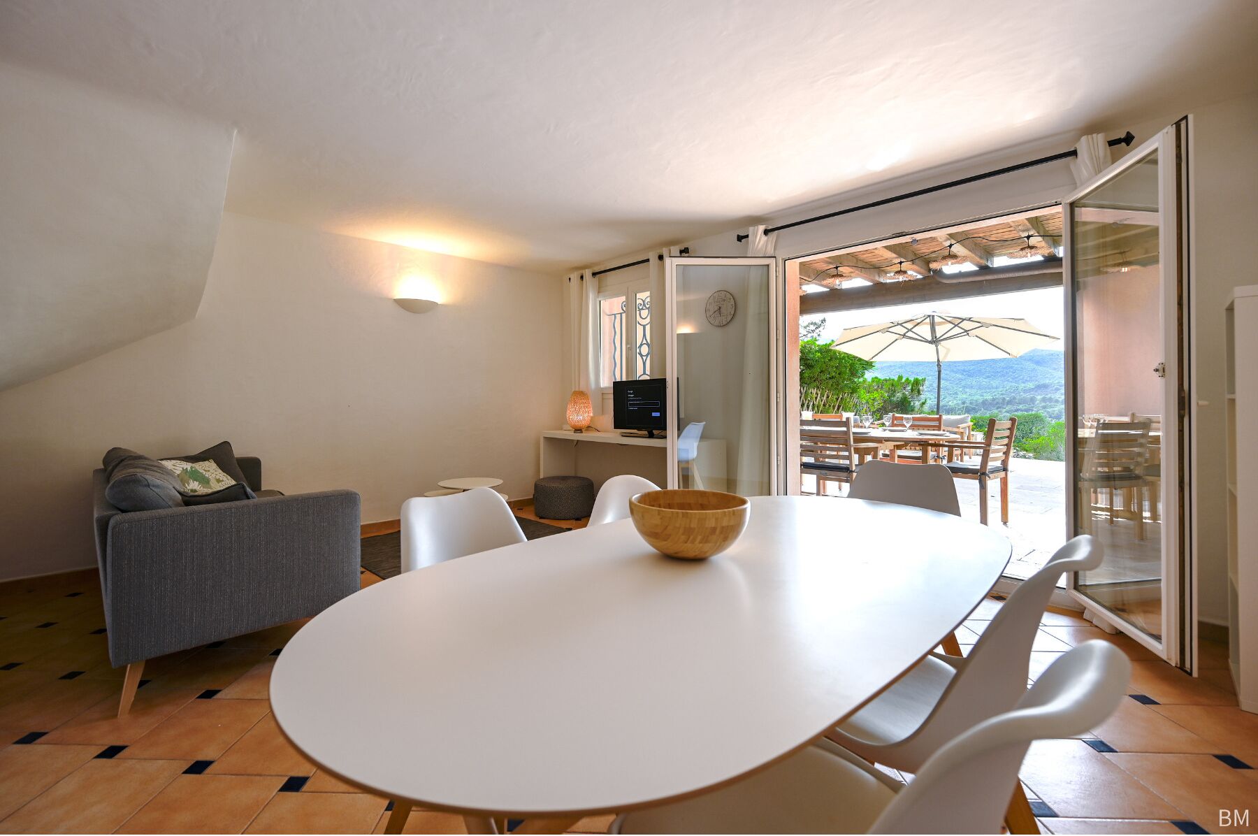 A modern dining area with a white table and chairs, a wooden bowl centerpiece, and a view of lush greenery through large sliding doors.