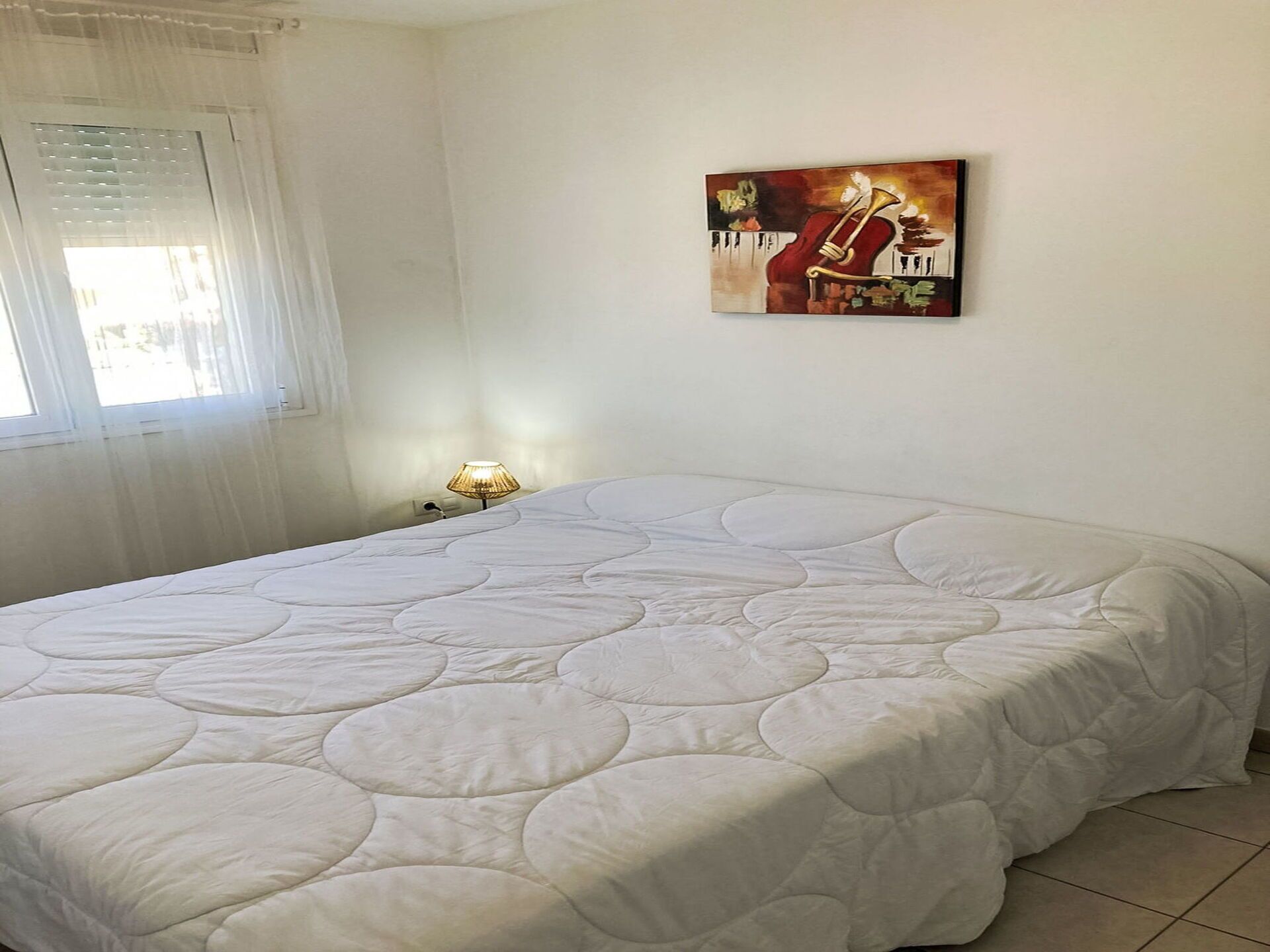 Ein Schlafzimmer mit einem großen Bett, einem Nachttisch mit Lampe, einem Fenster mit Jalousien und einem Bild an der Wand.