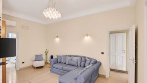Una sala de estar con un sofá modular, un televisor, una pequeña silla blanca y una araña.