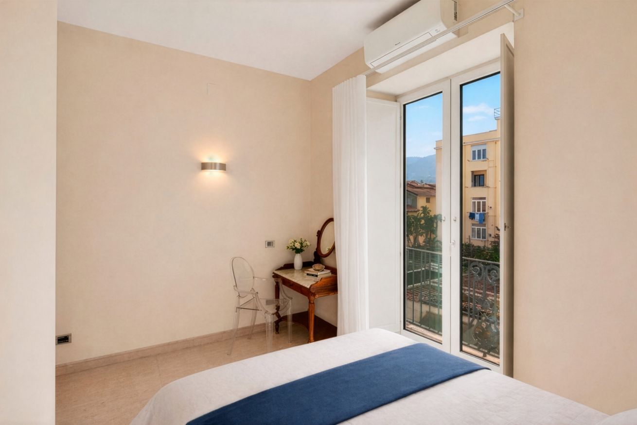 Habitación de hotel con una cama, un escritorio con una silla, una mesita con un florero y vistas a edificios a través de una puerta de cristal.