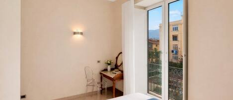 Habitación de hotel con una cama, un escritorio con una silla, una mesita con un florero y vistas a edificios a través de una puerta de cristal.