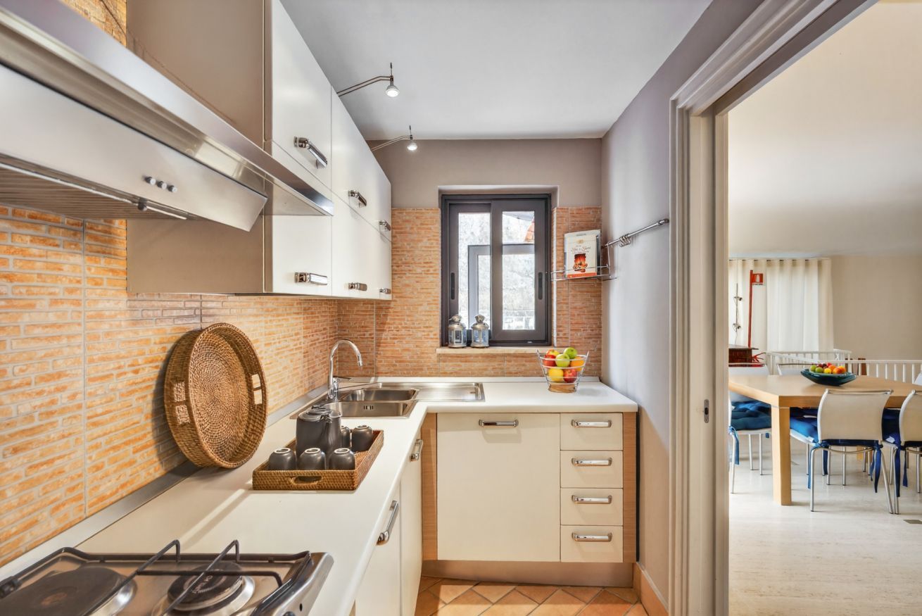 Cocina compacta con gabinetes blancos, estufa a gas, lavamanos y ventana con vistas.