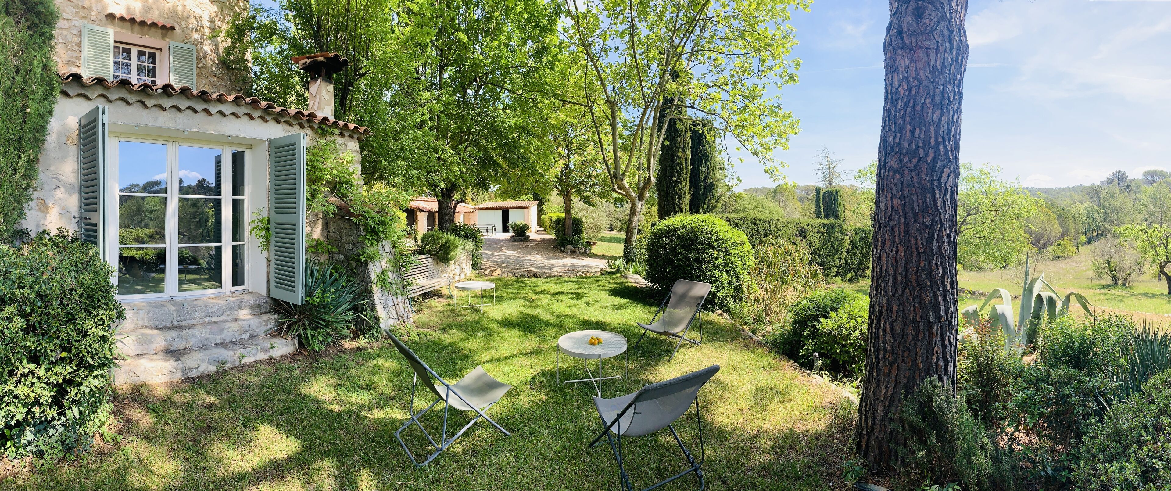 A rustic house with a stone facade, a small patio with a table and chairs, and a lush garden.