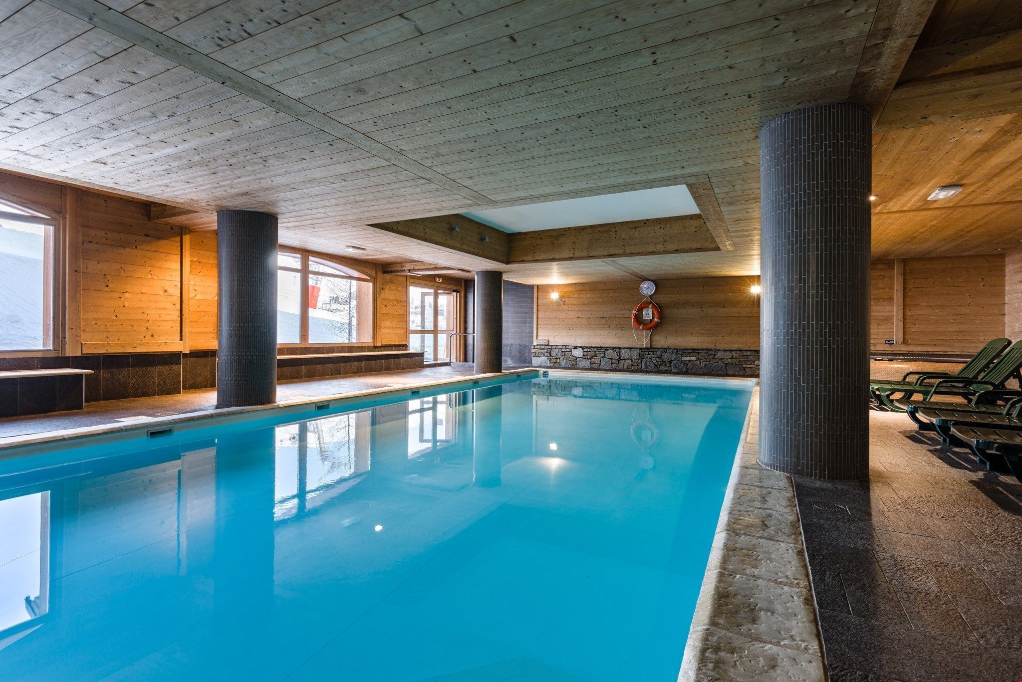 A large indoor swimming pool with wooden walls and ceiling, a lifeguard chair, and a stone wall with a fire extinguisher.