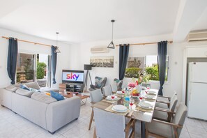 A modern living room with a sofa, dining table set for four, and a television displaying 'Sky'. - Villa Jazmin, Lovely 4BDR Protaras Villa with Pool,2 minute walk to Fig Tree Bay (Protaras)