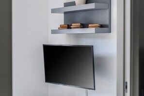 A modern interior with a flat-screen TV mounted on a wall, above a grey shelf with books and decorative items. - Modern Sorrento Apartment with Terrace&TennisCourt (Sorrento)