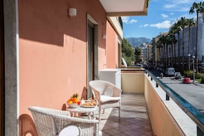 A balcony with wicker furniture, a table with fruits and pastries, and a view of a street with palm trees and buildings. - Modern Sorrento Apartment with Terrace&TennisCourt (Sorrento)