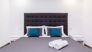 Ein ordentlich bezogenes Bett mit grauem Kopfteil, zwei blauen Kissen und einem weißen Handtuch daneben.