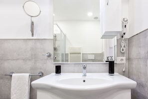 Un baño moderno con un lavamanos blanco, un espejo de mano y un secador de pelo en la pared.