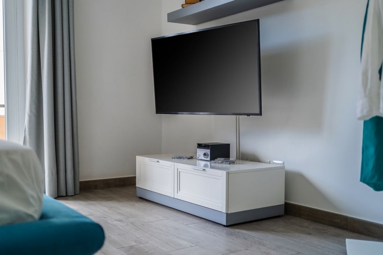 A modern room with a flat-screen TV mounted on the wall, a small cabinet with a radio, and a teal chair.